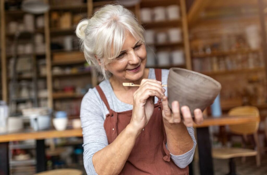Smiling senior woman carving a clay bowl in a pottery studio, wearing an apron and enjoying a hands-on craft activity.